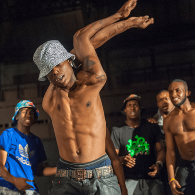 A shirtless dancer wearing a bucket hat entwines his arms over his head against a black background. Other dancers are visible behind him.