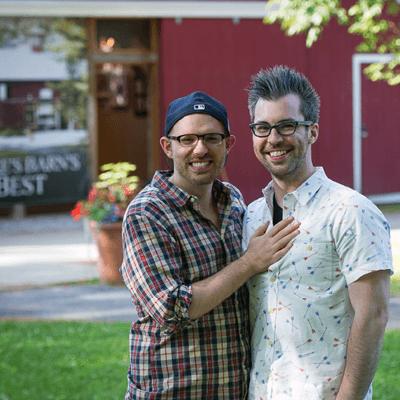 Two men stand in front with their arms around each other in front of a red barn.