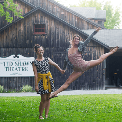 Two female dancers are in front of the Ted Shawn Theatre. The dancer on the left stands smiling at the camera and the dancer on the right is mid-leap with her arms extended back and leg extended forward.
