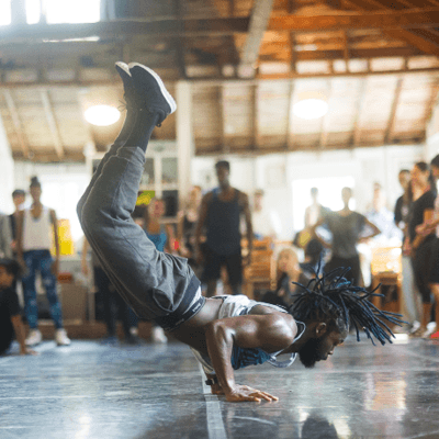 A male breakdancer holds himself up on his forearms with his legs in the air on an indoor stage. An audience is visible in the background.