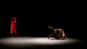 2 contemporary dancers cling to each other on the floor as a woman in a red dress sings behind them.