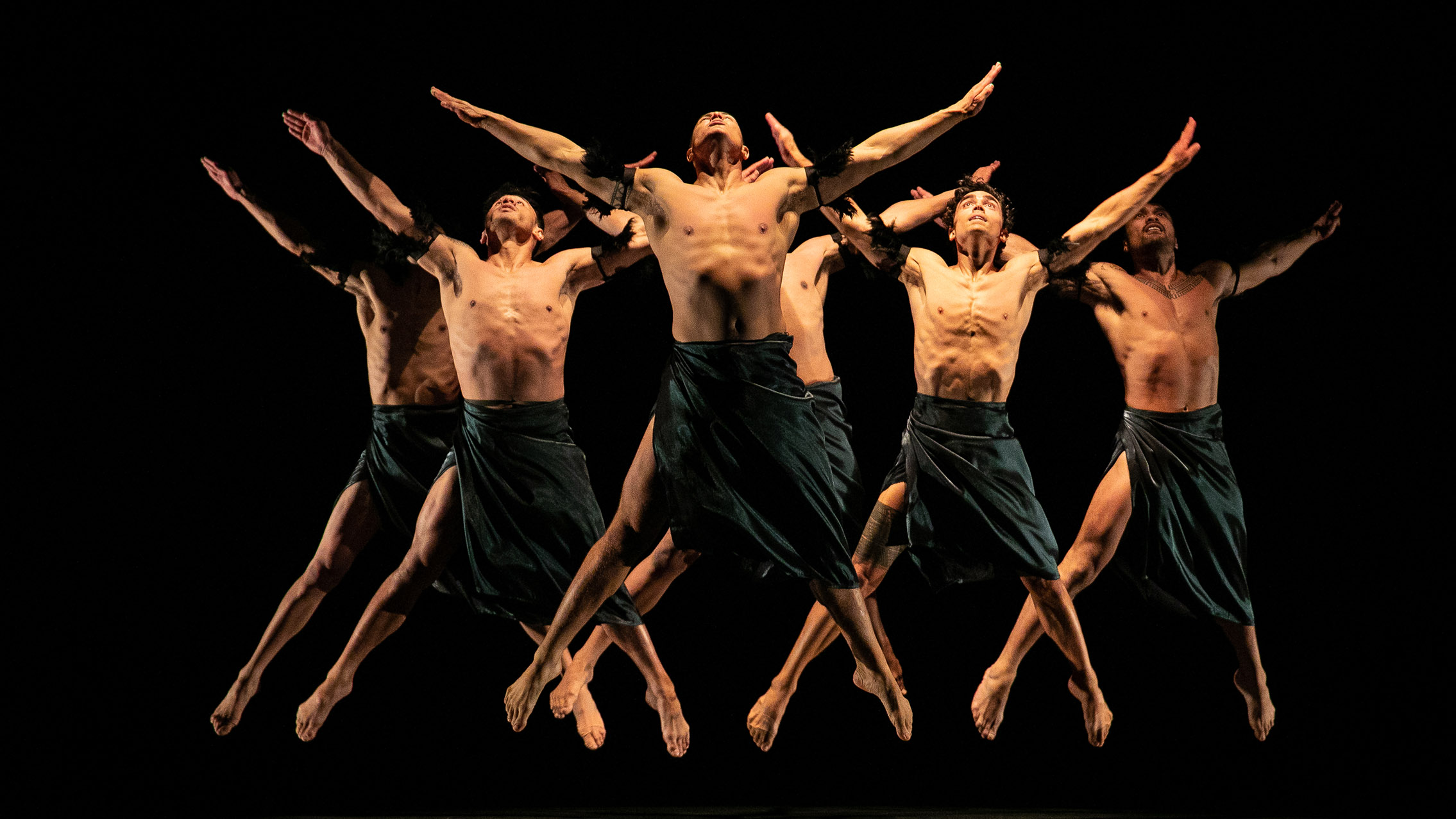A group of male presenting dancers leap in the air against a black backdrop.