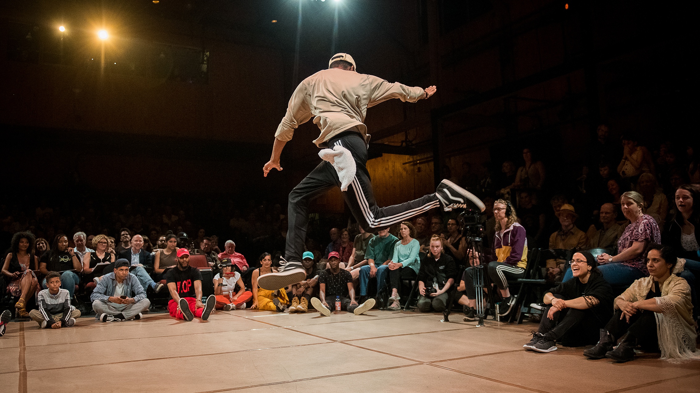 A dancer with his back to the camera is mid leap in an indoor studio. A seated audience is visible in the background.