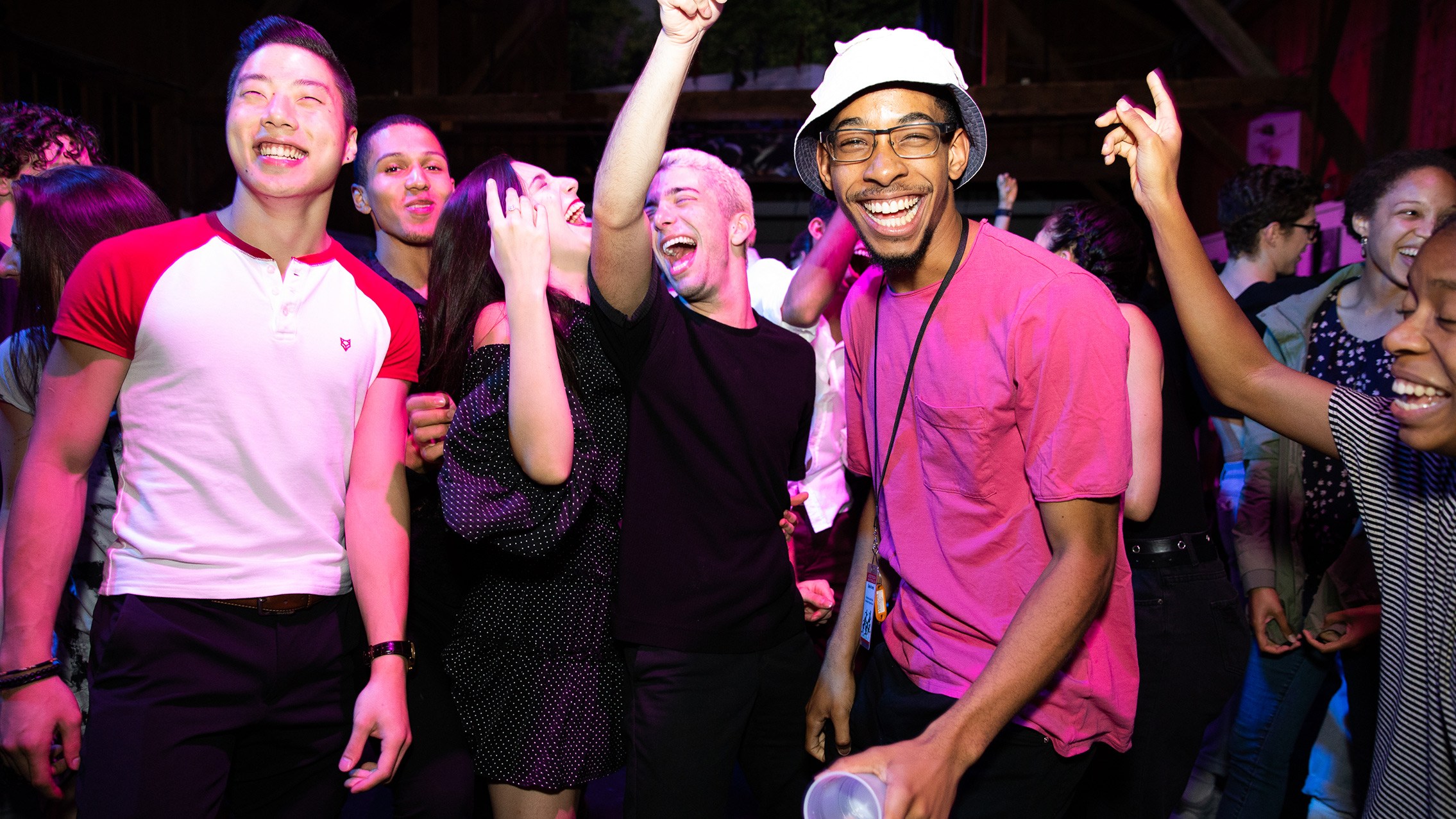 A group of people are smiling with their arms up under pink lighting.