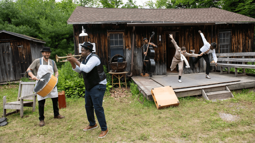 Two tap dancers are in sync on a platform in front of a cabin with an upright bass player right behind them. In front of them, to the left side, there is a trumpeter and drummer mid-playing.