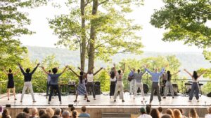 A zoomed out shot of a group of dancers with their backs to the audience and arms extended in V-shapes towards the sky. An audience is visible in the foreground and trees are visible in the background.
