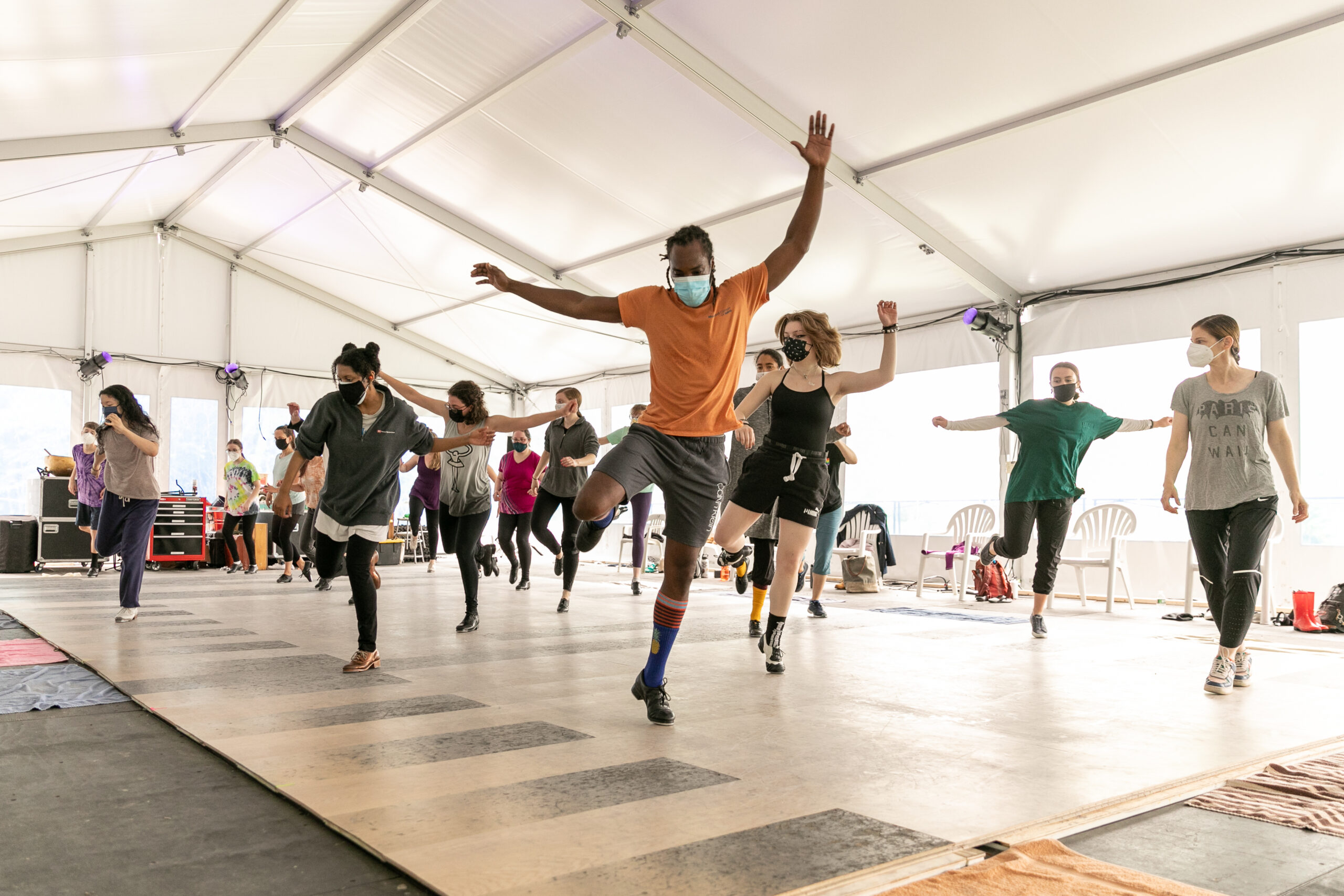 A group of tap dancers take class in an outdoor tent. They all are kicking their right foot and raise their arms in various shapes.