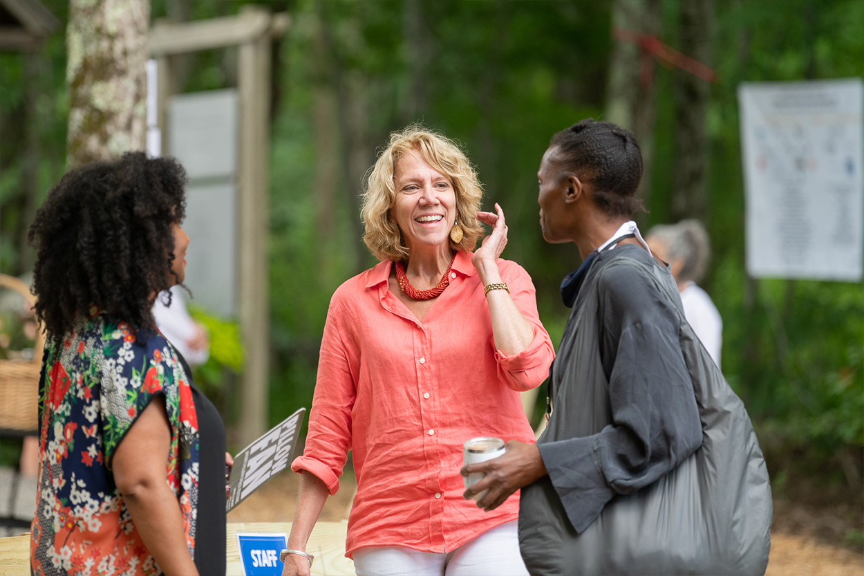 Outside stands three women visible from the waist up. Melanie, a black-skinned woman with short, coily black hair who is wearing a multicolored blouse is on the left. She is looking at Pamela, a white woman with shoulder length blonde wavy hair and a coral button up top. She is smiling and looking at Okwui on the right, who is a black-skinned woman with short black hair. Okwui is wearing a gray blouse and has a tote bag on her shoulder.