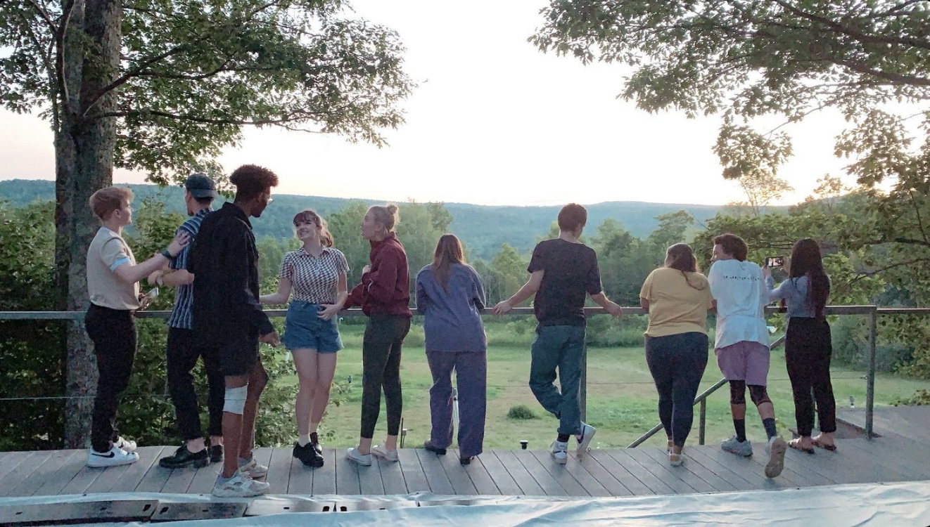A group of people stand on an outdoor stage facing away from the camera and overlook the hills and trees beyond.