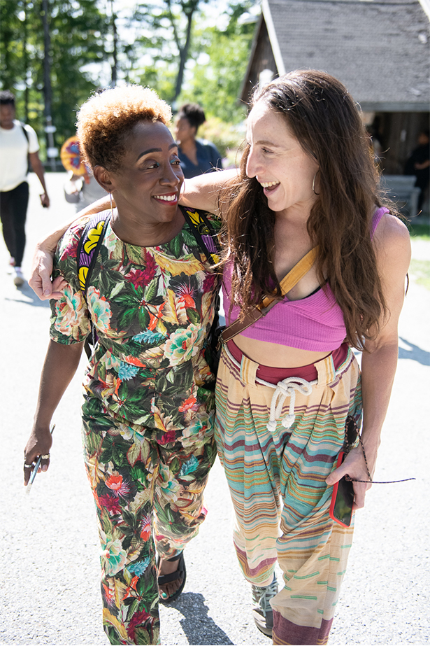 Two woman are smiling, walking together outside. On the left is LaTasha, a black skinned woman with tight coiled curly hair who is wearing a floral patterned jumpsuit. She is looking at the woman on the right, Ephrat, a white woman with long brown hair. Ephrat is wearing a pink crop top and multi-colored striped pants and has her arm around LaTasha's shoulders.