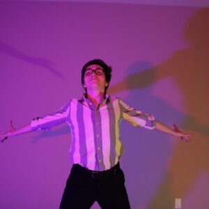 A dancer throws his arms open facing the camera. He wears a striped shirt and black pants. The lighting is purple and his figure casts a shadow on the wall behind him.