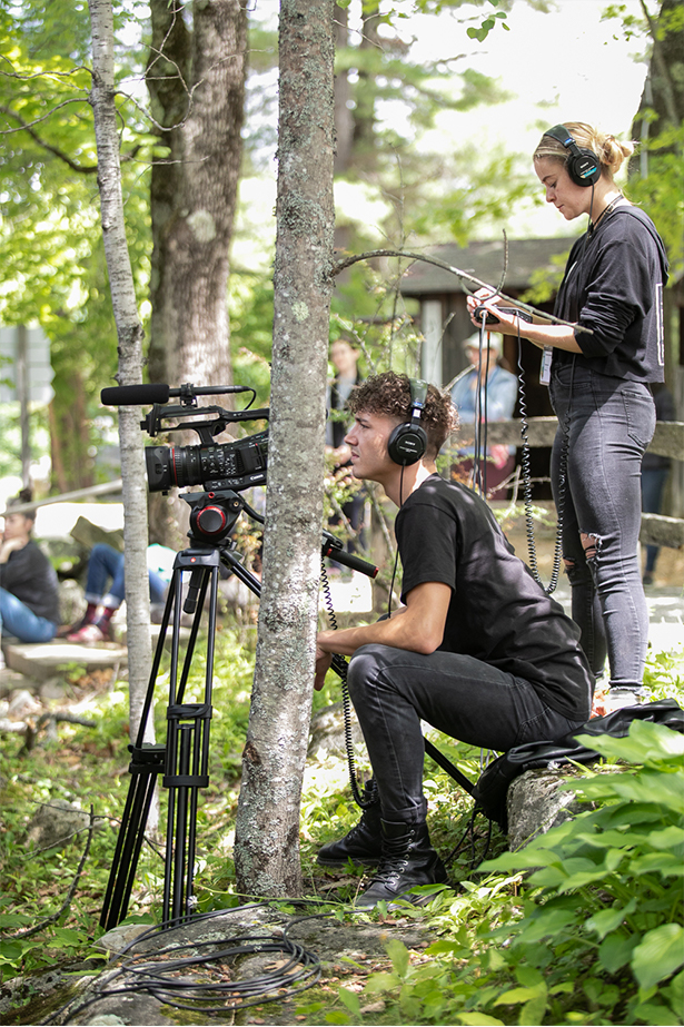 Outside amongst the tress are two members of the video team. Mason, a white man with curly brown hair, sits wearing all black and operating a video camera. Sydney, a white woman with blonde hair in a low bun, stands behind him wearing all black and a headset.