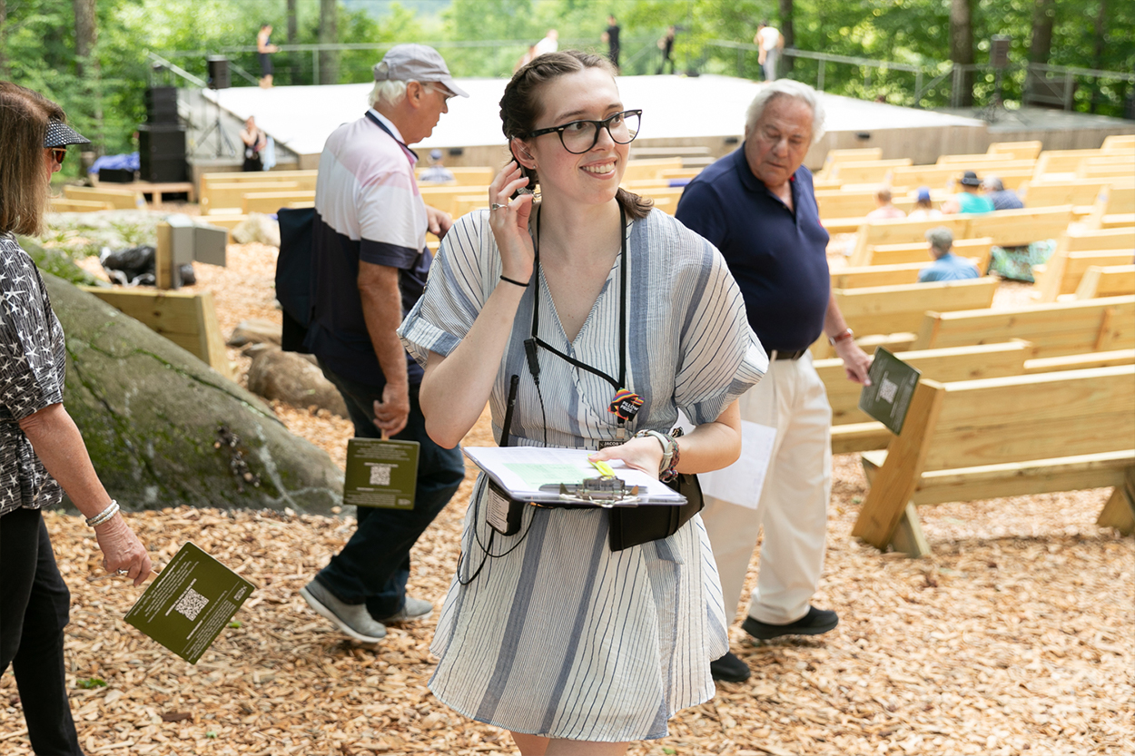 Brenna, a white woman with brown hair in braided pig-tails stands outside wearing a white and blue striped dress and glasses. She's holding a clipboard and smiling while looking at someone out of frame. Behind her are wooden benches facing an outdoor stage.