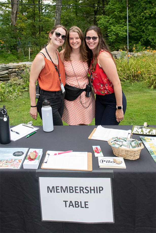 Three woman stand together smiling outside in front of a table labeled "Membership Table." On the left is Kristen, a white woman with brown hair wearing an orange tank top, black pants, and sunglasses. In the center is Taylor, a white woman with long dirty blonde hair wearing a pink and white polka-dot dress. And on the right is Alexis, a white woman with long dark hair wearing sunglasses, a red tank top and navy pants.