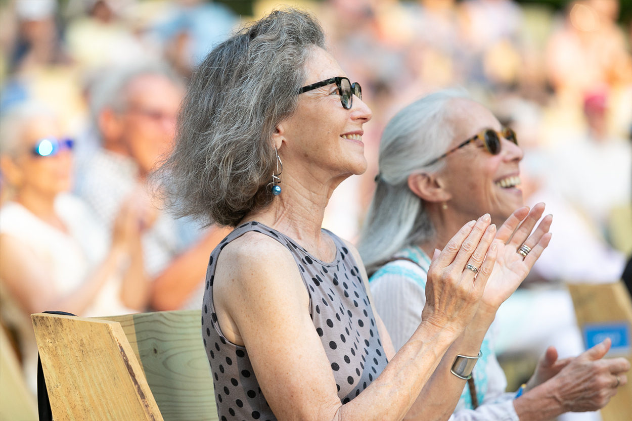 Two women sit beside each other outside only visible from the waist up. They are in profile, each captured with their hands clapping mid-applause and with full, joyous smiles on their face. The woman on the left has shoulder length, dark gray hair and is wearing black framed glasses and a gray tank top with black polka-dots. The woman on the left has long, white hair and is wearing a white and blue blouse.