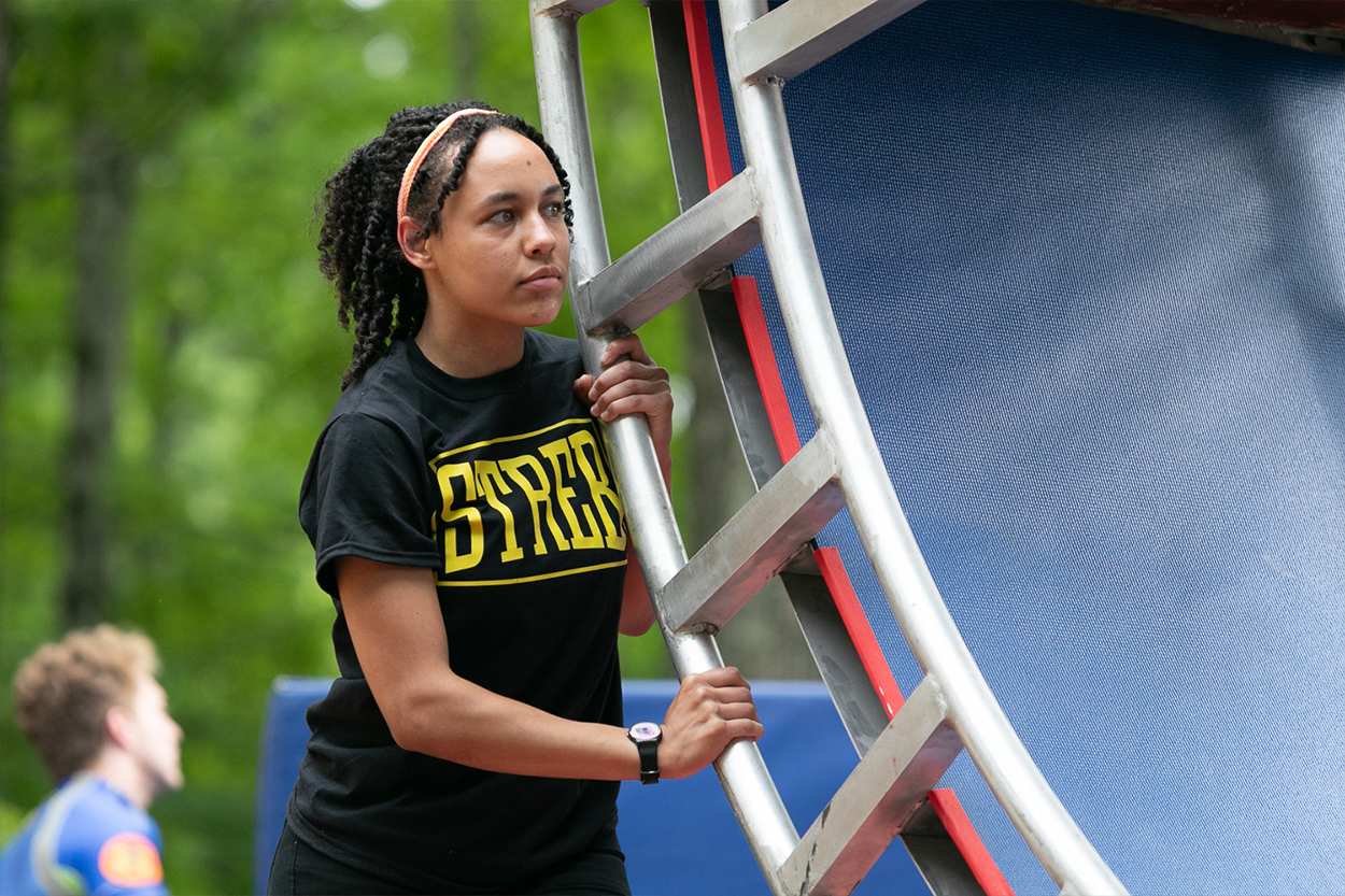 Erin, a brown skinned woman with coily black hair is wearing a black tee shirt that says "STREB" in all capital yellow letters. She is outside and holding onto a curved metal bar.