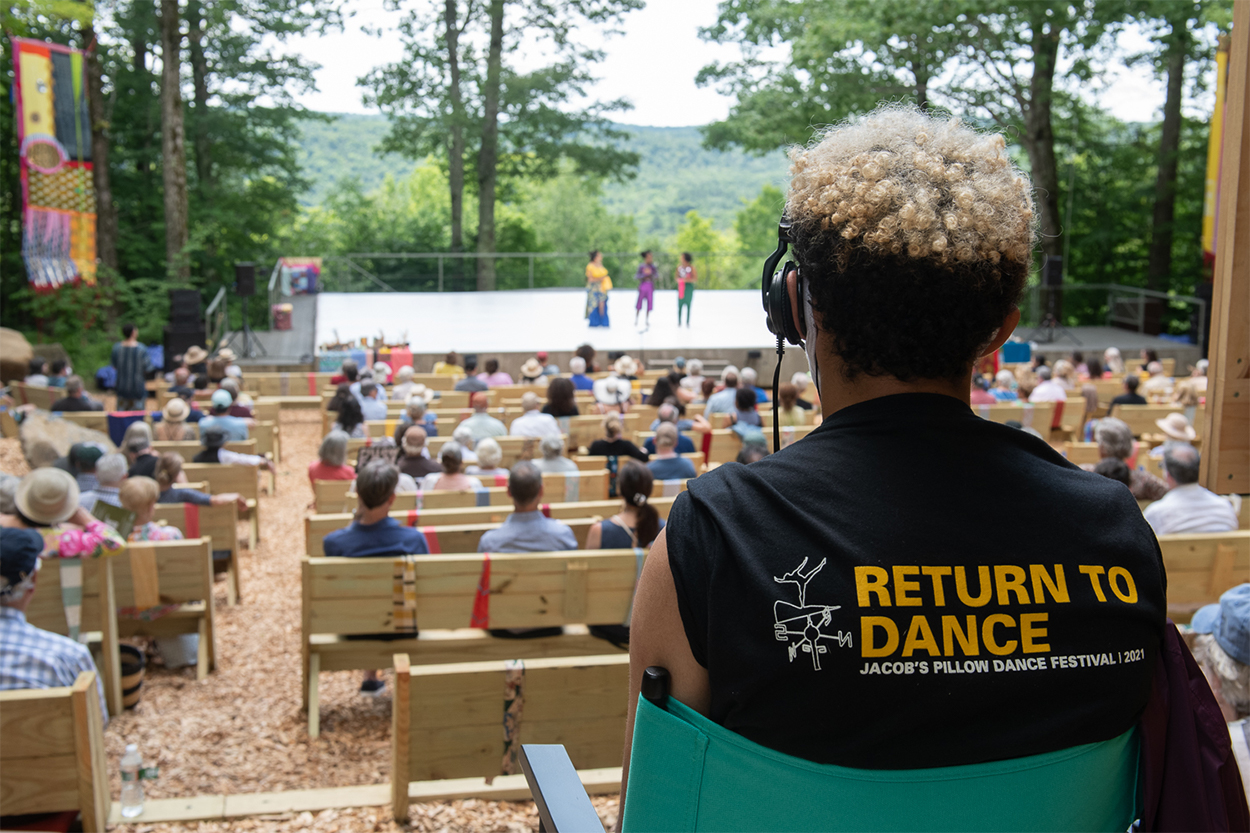On the right is Lex, a brown skinned man with short, tight black curly hair with a circular patch of blonde on top. Seen from behind, he sits in a director's chair and is wearing a black tee shirt. Across the back in bold, yellow, capital letters it says "Return to Dance," and in small white capital letters underneath it says "Jacob's Pillow Dance Festival 2021." In front of Lex is a full audience sitting outside on wooden benches looking at a stage surrounded by trees.