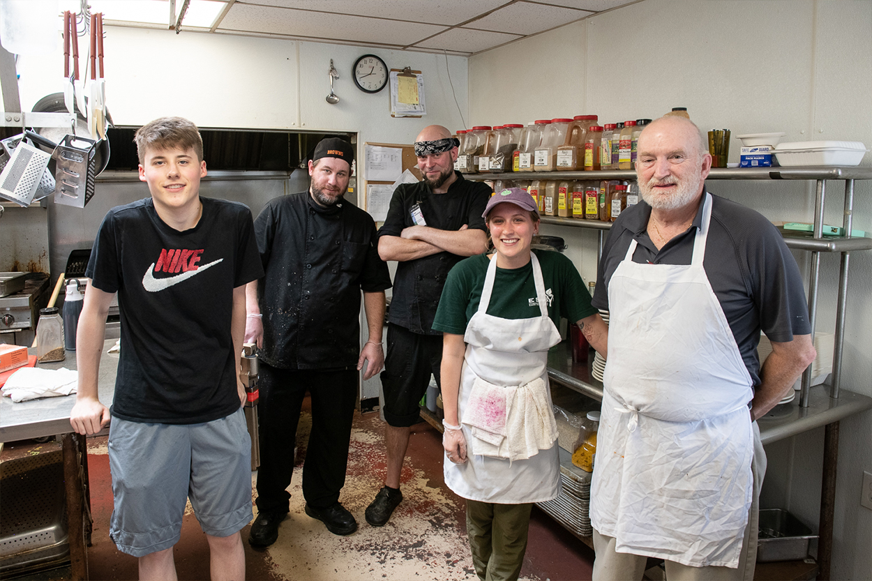 Standing in a line are five dining staff members. Three men on the left, then a woman, and another man on the right. They are standing in a kitchen with shelves of cookware and spices behind them.