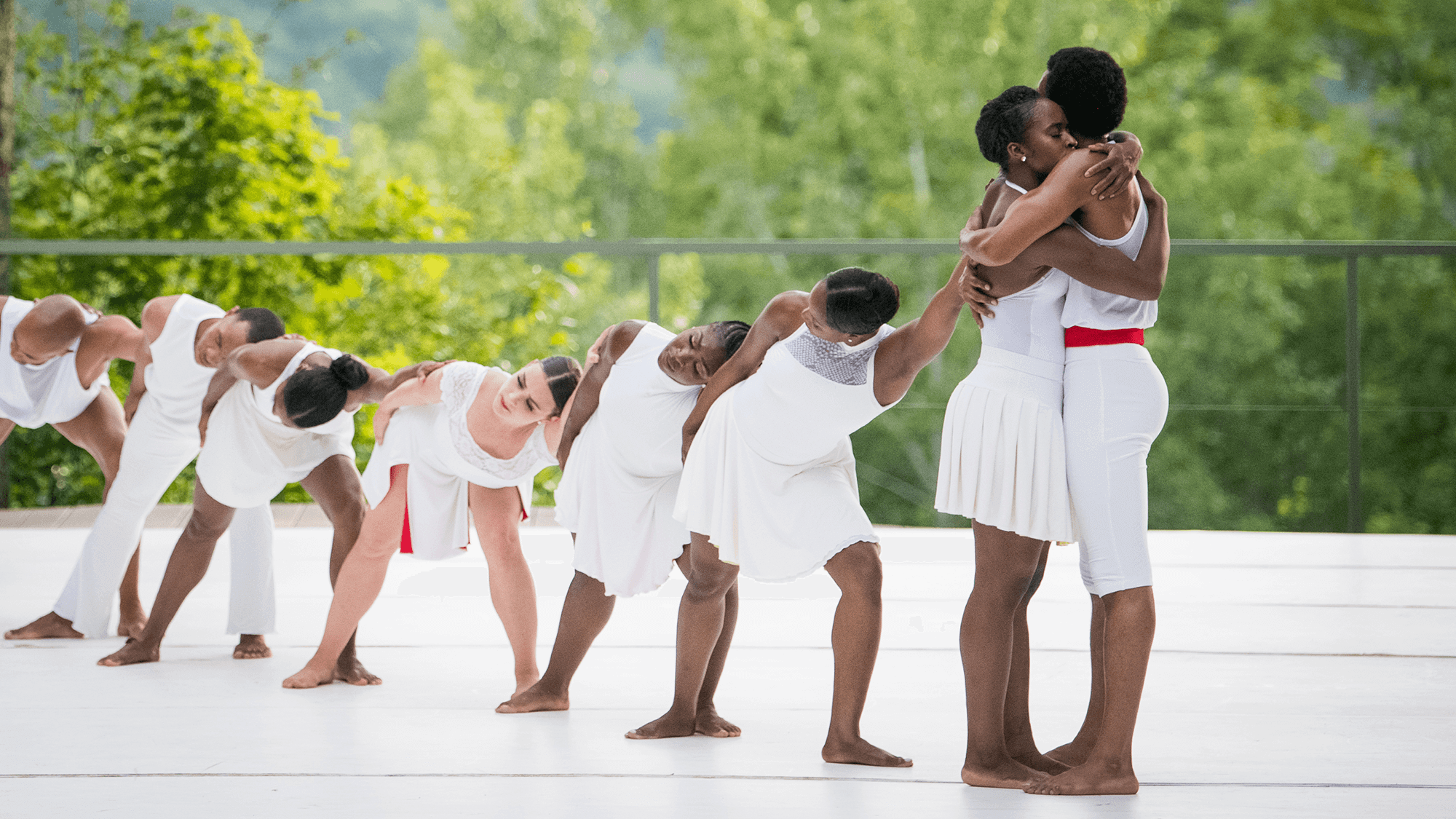 Two dancers wearing white embrace while six dancers in white behind them lunge forward in a line with their left arms outstretched. Trees are visible behind them.