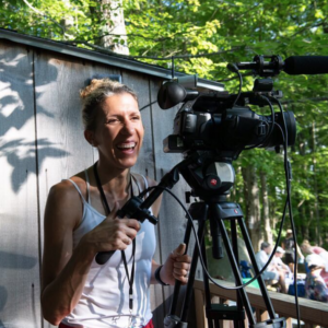 Videographer Nel Shelby stands at the back of the outdoor Henry J. Leir Stage amid leafy green trees, laughing while controlling a video camera on a tripod.