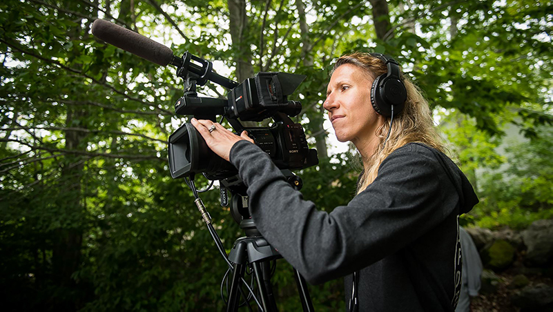 Videographer Nel Shelby stands in the forest wearing headphones, looking through a video camera on a tripod