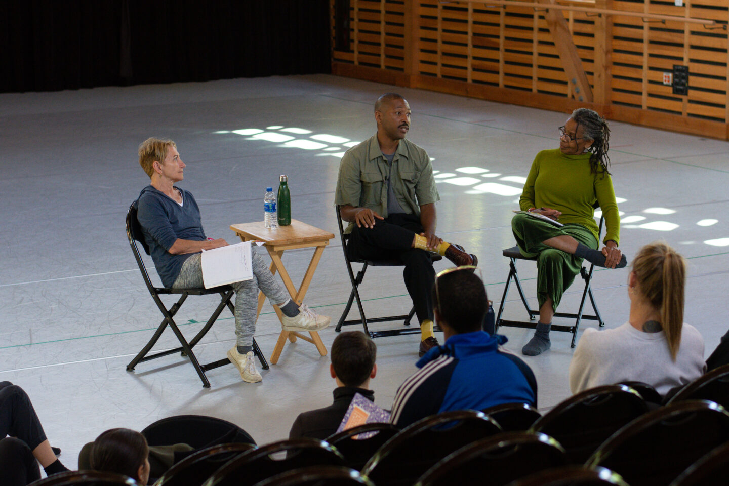 Program Directors Risa Steinberg and Bebe Miller sit on either side of Guest Speaker Kyle Abraham in a studio. Facing them is a group of people sitting in chairs and watching.