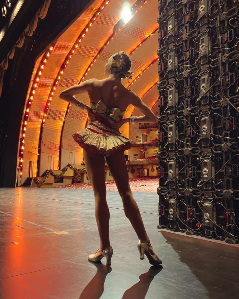Kathleen Laituri stands with her back to the camera. Her hands are on her hips as she looks upward and extends her left leg long. She faces the empty Radio City Music Hall.