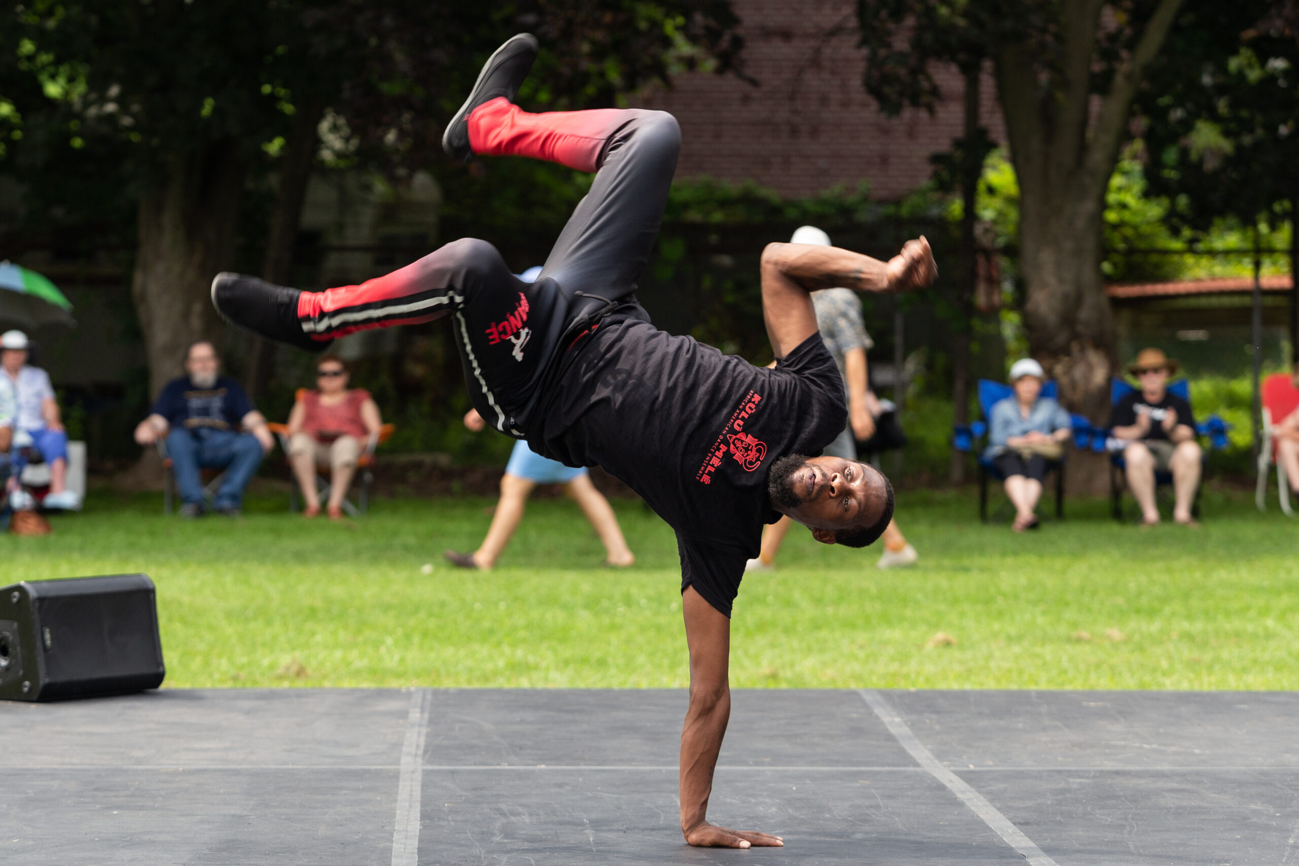 A dancer in a one-handed handstand with arms and legs bent. The dancer is on an outdoor stage and there are onlookers in the background.