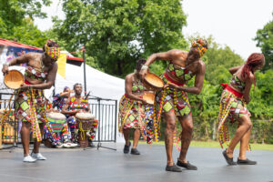 Four dancers stand on an outdoor stage wearing colorful prints and holding small drums.