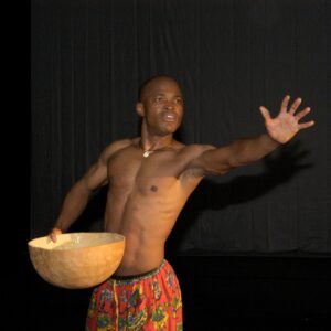 Iddrisu Saaka stands facing the camera against a black background. He is shirtless and wears multicolored pants. He extends his left arm, spreading the palm of his hand toward the camera. In his right hand, he holds a bowl.