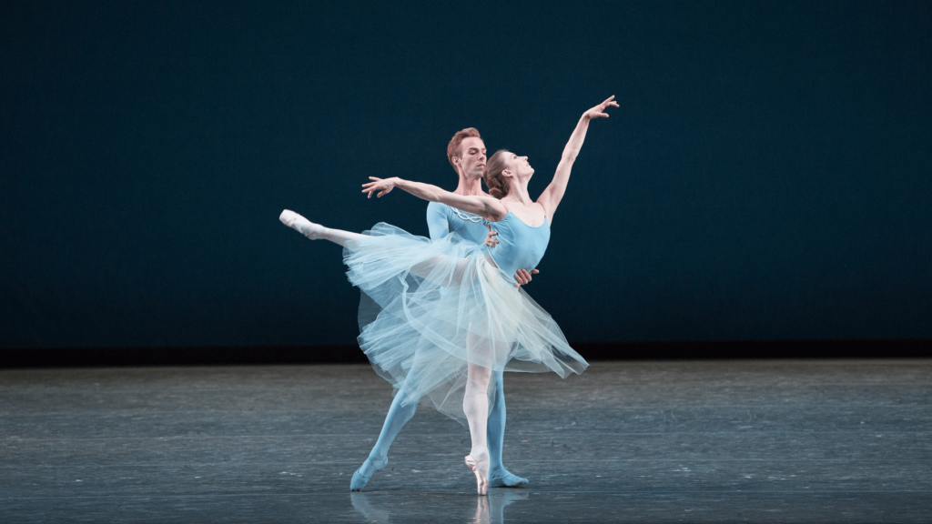 A pair of dancers placed in front of each other in light blue ballet attire one in tights and the other wearing a loose tutu. The dancer in the back supports the dancer in front who is balancing on pointe with a leg extended behind. Photo by Alexander Iziliaev