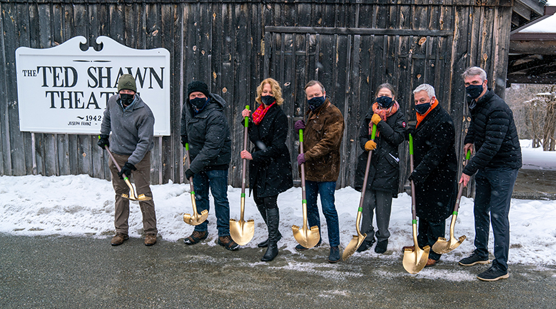Seven people wearing masks and winter clothing stand holding golden shovels outside of the Ted Shawn Theater.