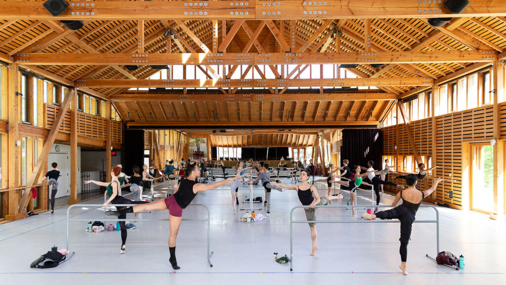 Dancers stand at barres in a large studio in class in an arabesque position, with their left legs extended back at a 90 degree angle and their right arms reaching forward.