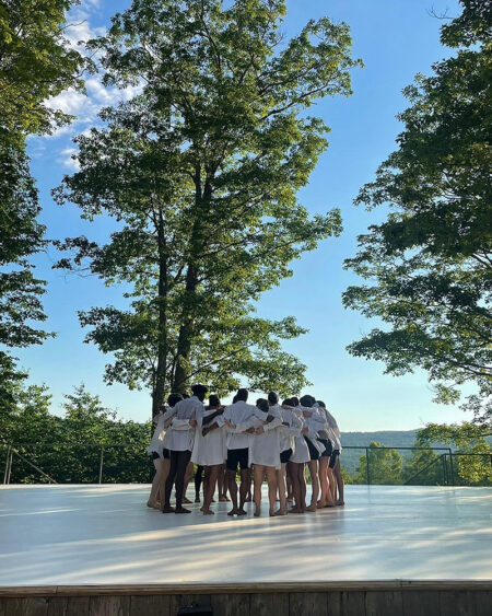 A group of dancers wearing white shirts and black shorts embrace on an outdoor stage.