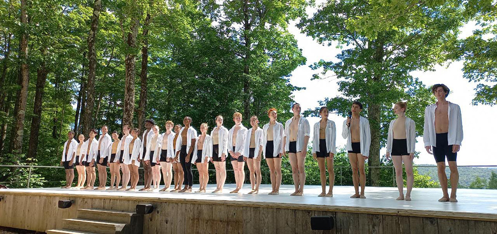 Dancers wearing white shirts and black shorts stand in a line on an outdoor stage as if about to bow.