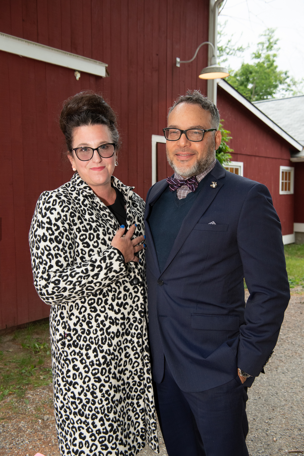 A man and woman pose for the camera. He is wearing a navy suit jacket and she is wearing a leopard print coat.