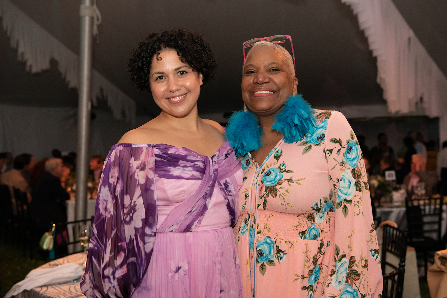 2 women smile for the camera, wearing purple and pink dresses.