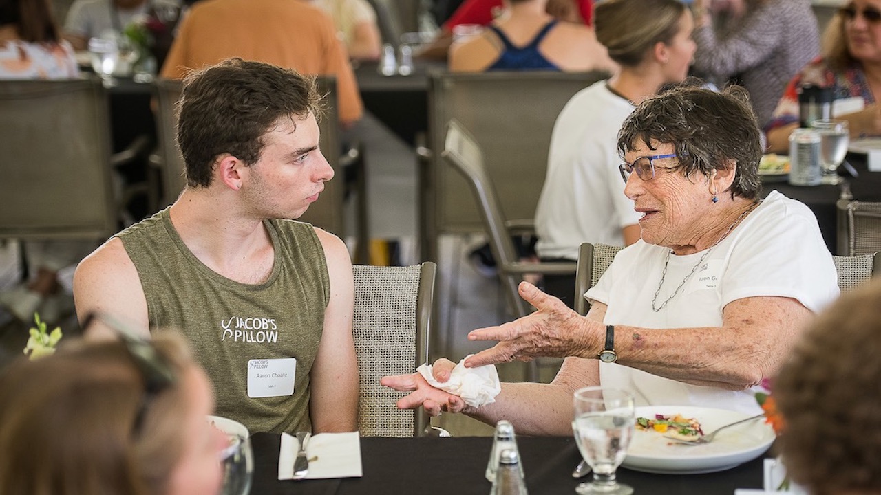 A young white man listens to an elderly white woman speak at lunch.