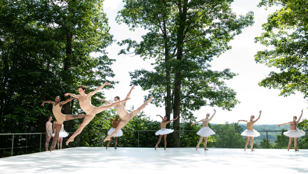Three shirtless dancers leap in the front of an outdoor stage while dancers in tutus stand in the back.
