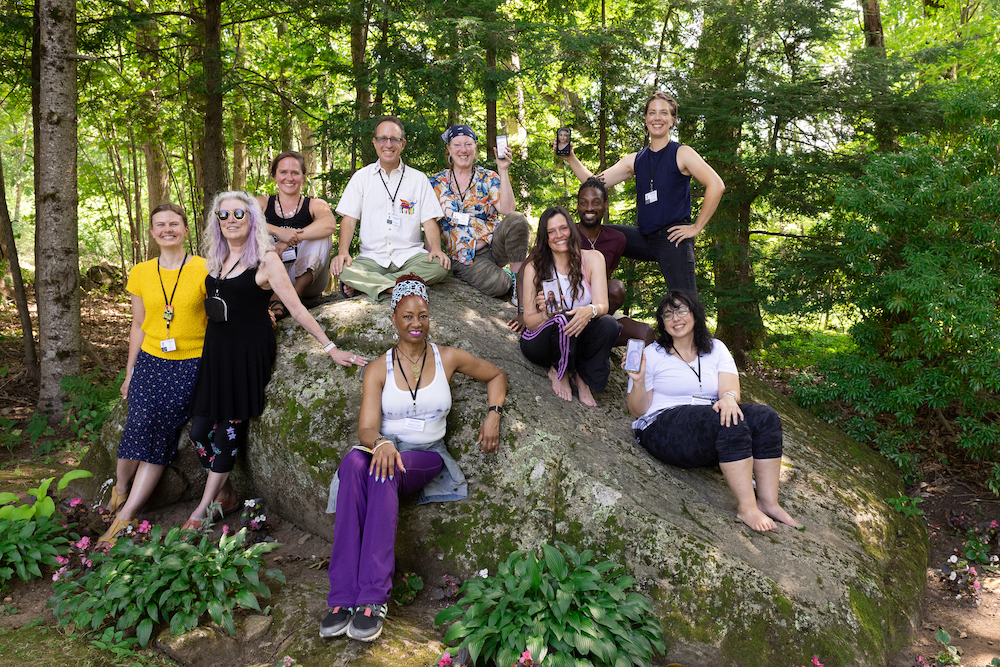 10 individuals in the 2021-2022 Jacob's Pillow Curriculum in Motion Institute cohort pose on the Pillow Rock. 4 members are holding up their phones to show members participating remotely.