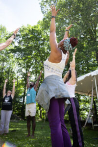 Members of the 2021-2022 Curriculum in Motion Institute stand in a circle and raise their arms towards the sky during a session. They are outside standing in a lawn and under trees.