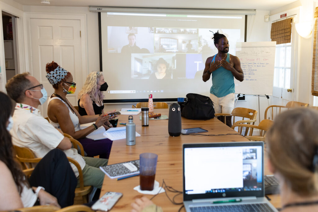 Members of the 2021-2022 Curriculum in Motion Institute sit around a wood table while one members stands and speaks to them. Behind this member is a screen, where members participating remotely are on Zoom.