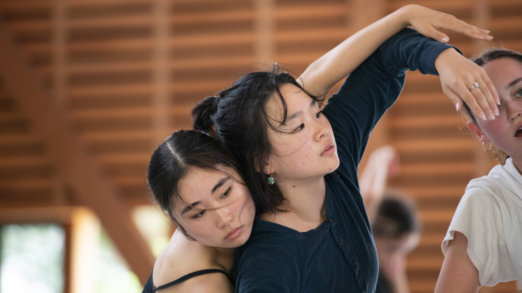 Two dancers move together, one in the back with arms draping over the arms of the dancer in the front.