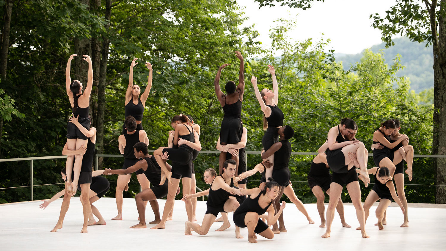 A group of dancers wearing black assume various poses from high to low levels on an outdoor stage.