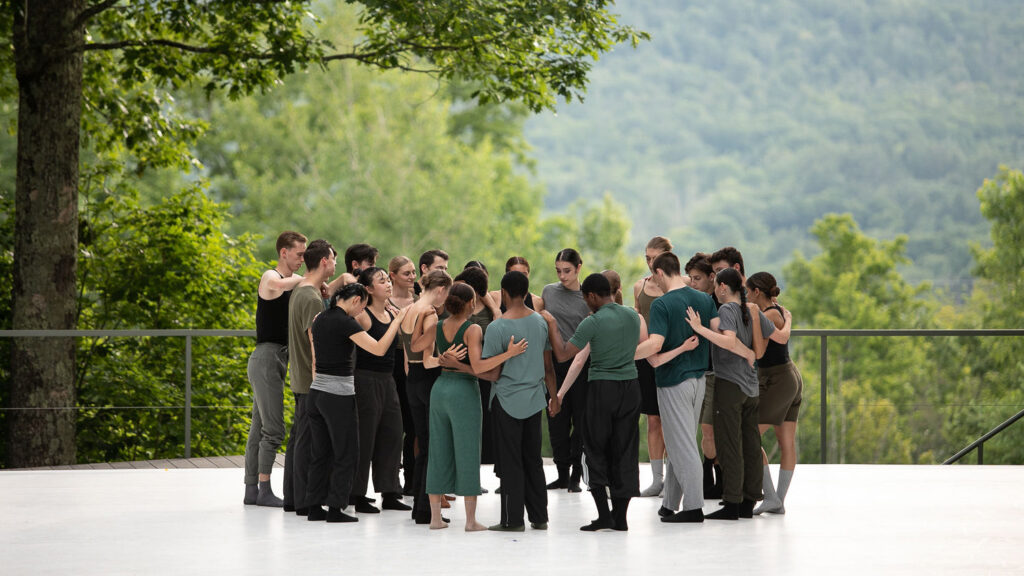 A group of dancers wearing varying shades of green, grey, and black embrace in a large circle on an outdoor stage.