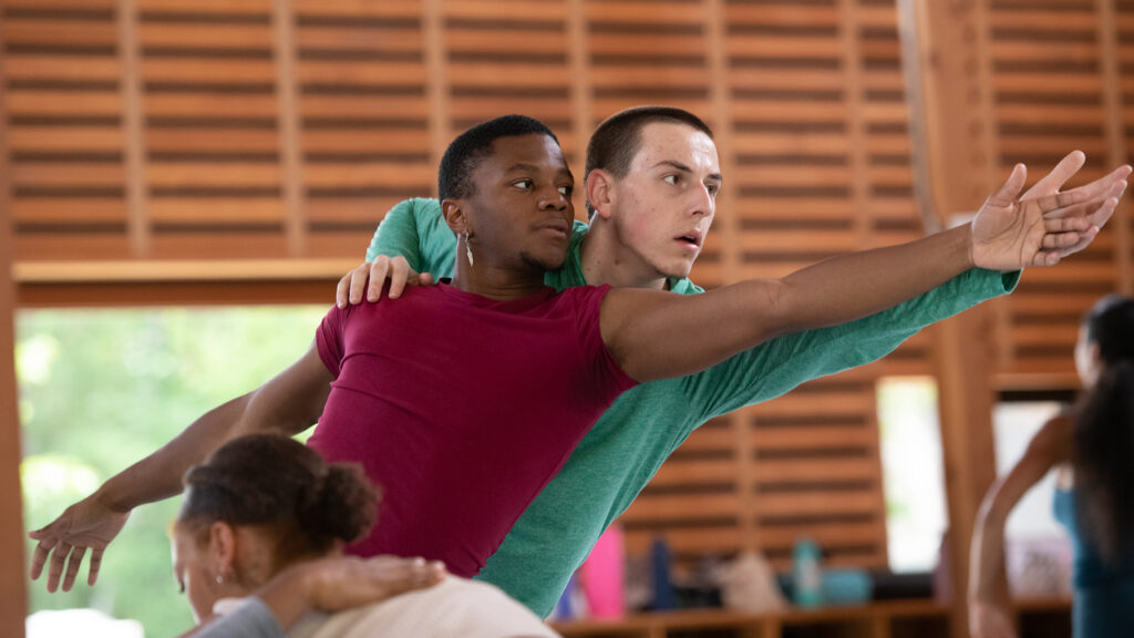 Two dancers reach with their left hands. The dancer behind places a hand on the other dancer's left shoulder. Both look and lean to the left.