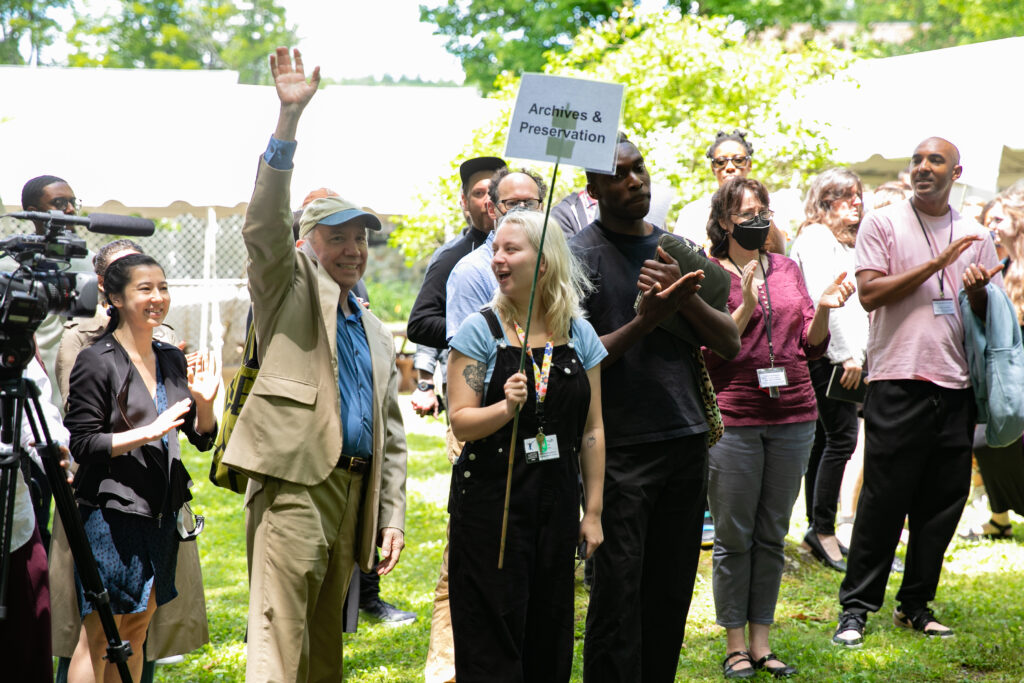 A group of people smile and clap as intern Hannah Berry holds a sign that says "Archives & Preservation" and Norton Owen holds up his hand.