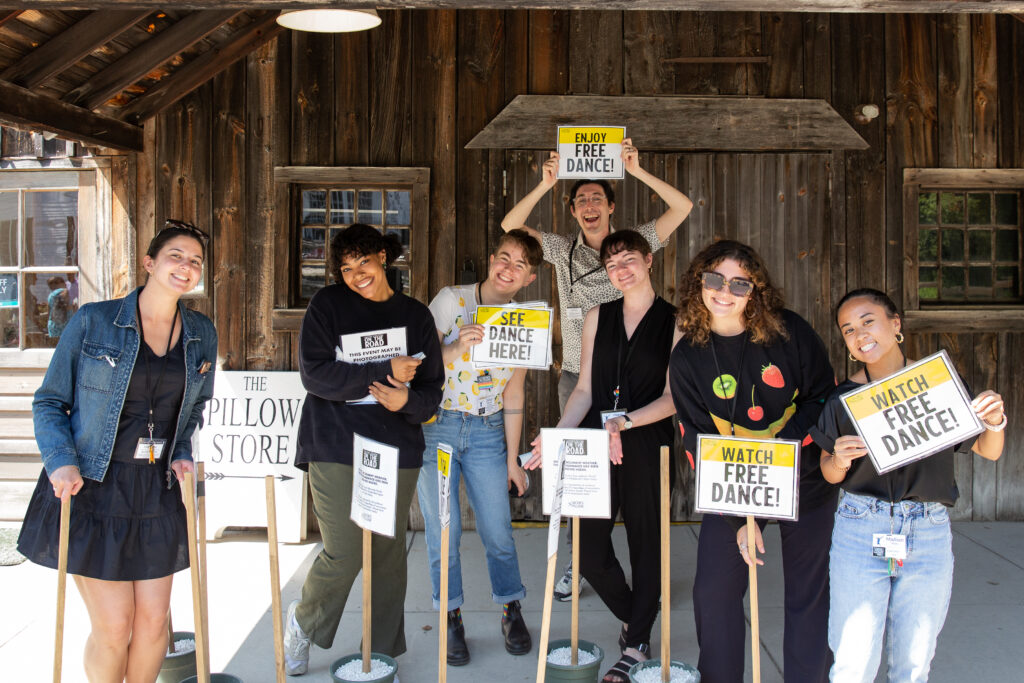 7 individuals smile and hold up signs that say "See Dance Here!" and "Watch Free Dance!"