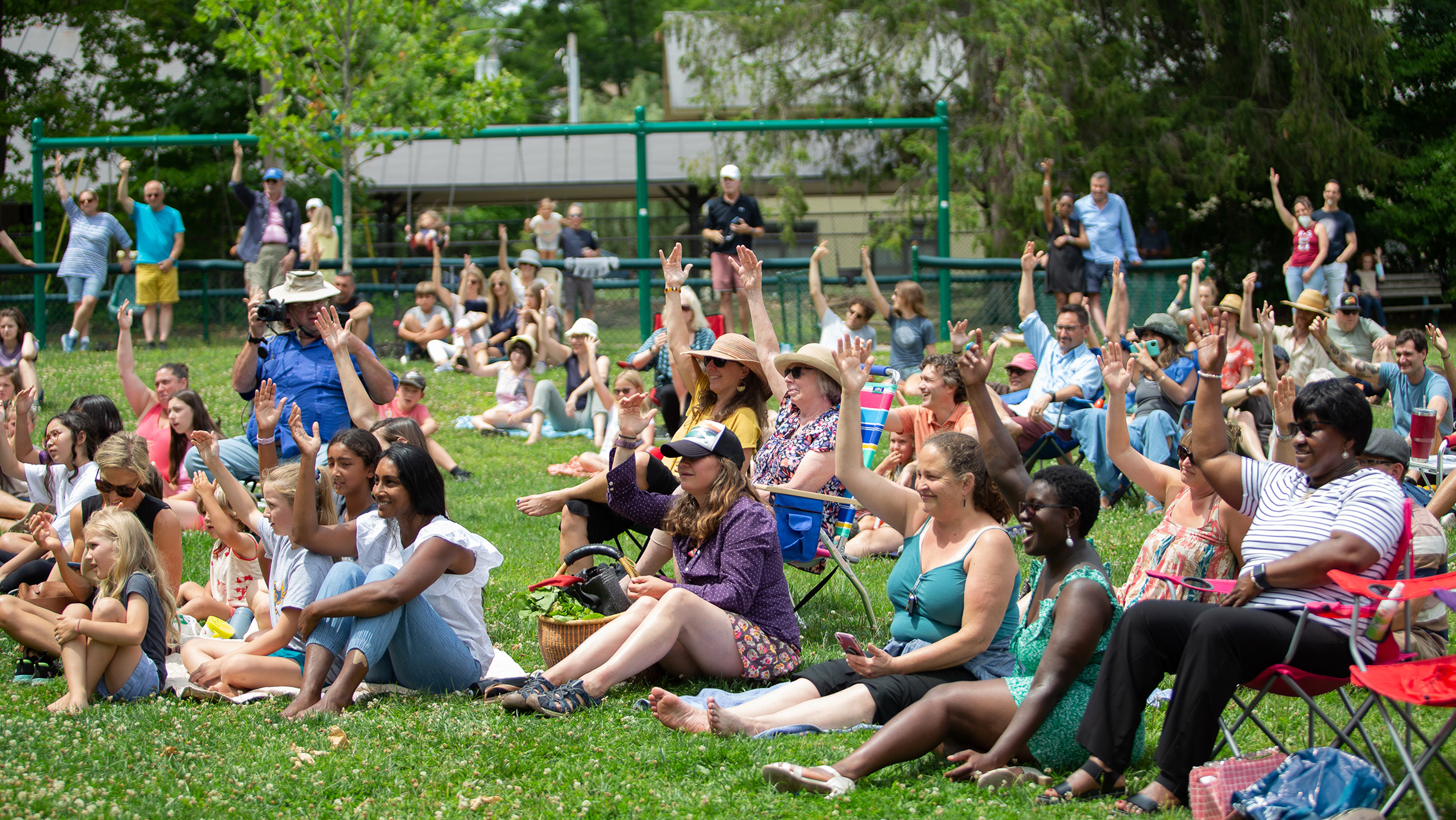 A group of people in a grassy park, with some sitting in chairs, sitting on the grass, or standing. They raise their arms.