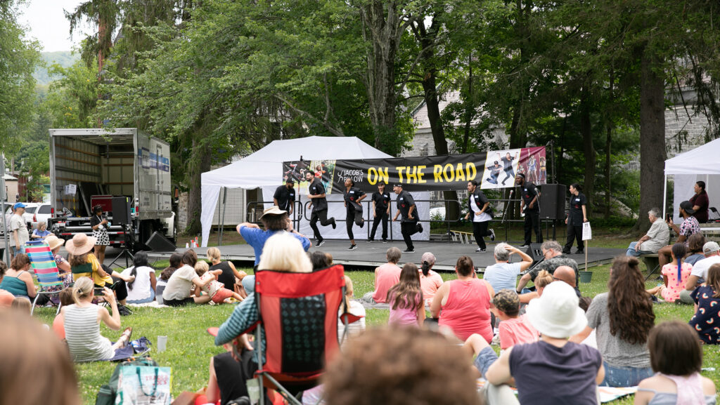 A group of people sitting in a park face an outdoor stage with a sign that says "On the Road" behind it. The dancers are dressed in all black.
